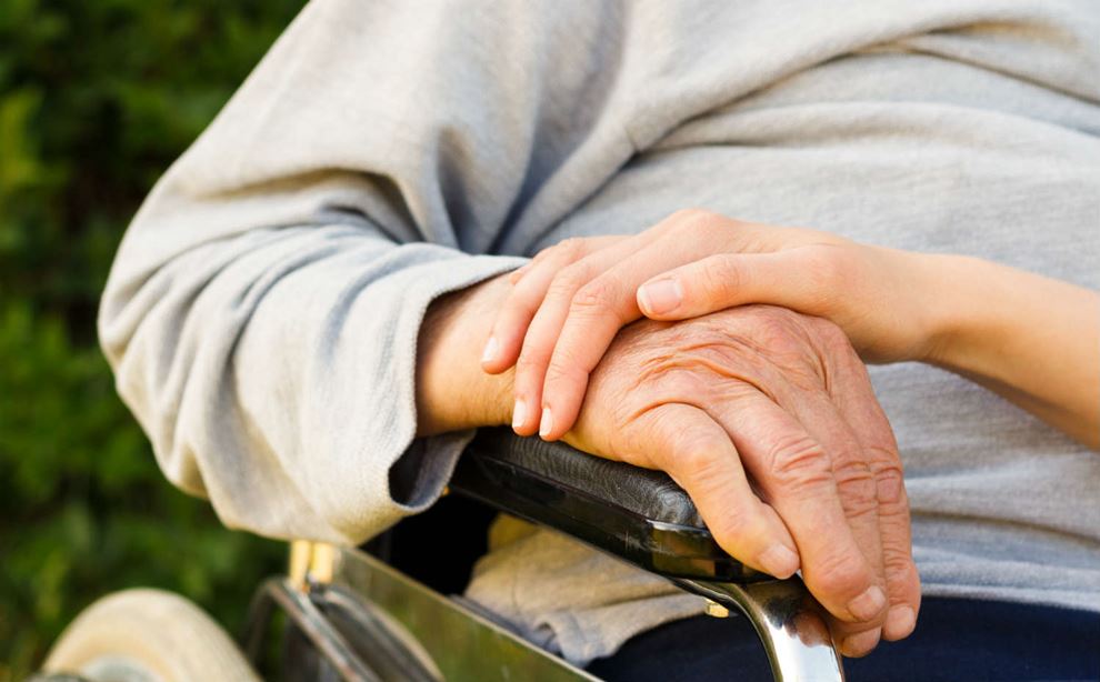 En yngre hånd holder på en ældre hånd, som hviler på et armlæn på en kørestol